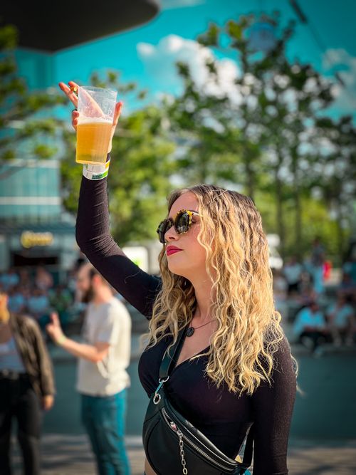 Free Blonde woman enjoying a sunny day outdoors with a glass of beer, surrounded by people. Stock Photo