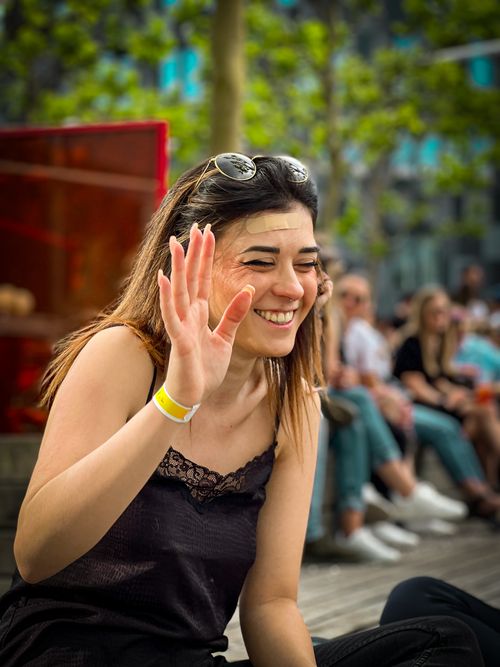 Free Joyful woman in casual attire, smiling with hand raised, enjoying a sunny outdoor setting. Stock Photo