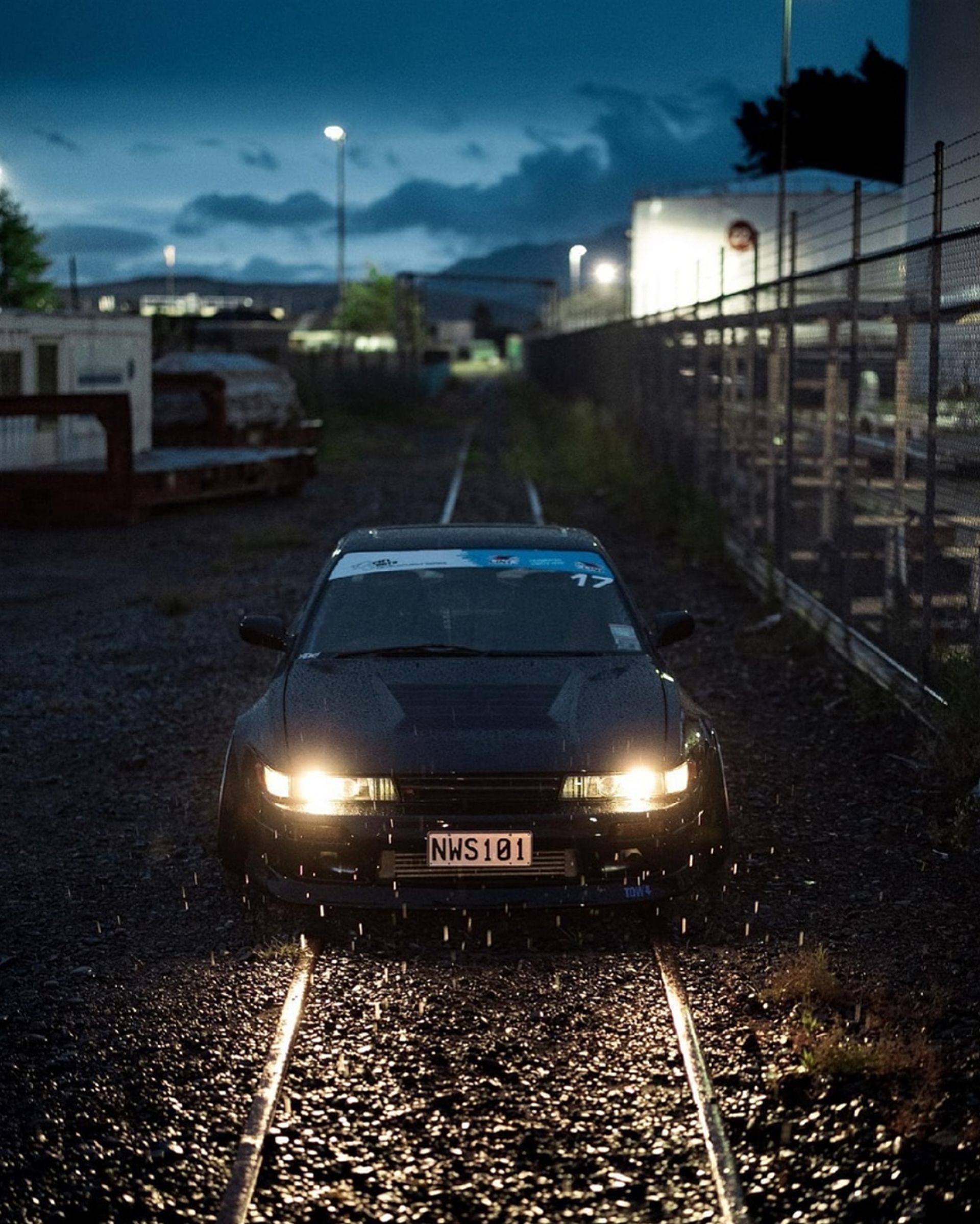 Black vintage car with headlights on, parked on rainy railway tracks at night.