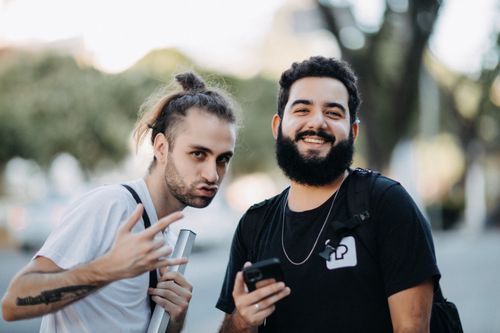 Free Two young men outdoors, smiling and holding cellphones, in a casual setting. Stock Photo