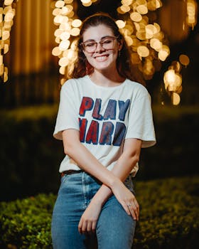 Young woman smiling warmly in stylish glasses and casual 'Play Hard' shirt, outside with decorative lights.