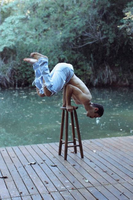 A shirtless man balances acrobatically on a stool by a tranquil riverside deck.
