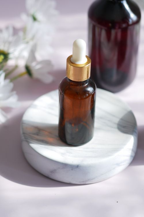 Free Brown glass dropper bottle on a marble platform with flowers, ideal for beauty product mockups. Stock Photo