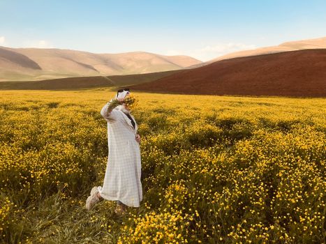 Woman standing in a yellow flower field with hills in the background, obscured face.