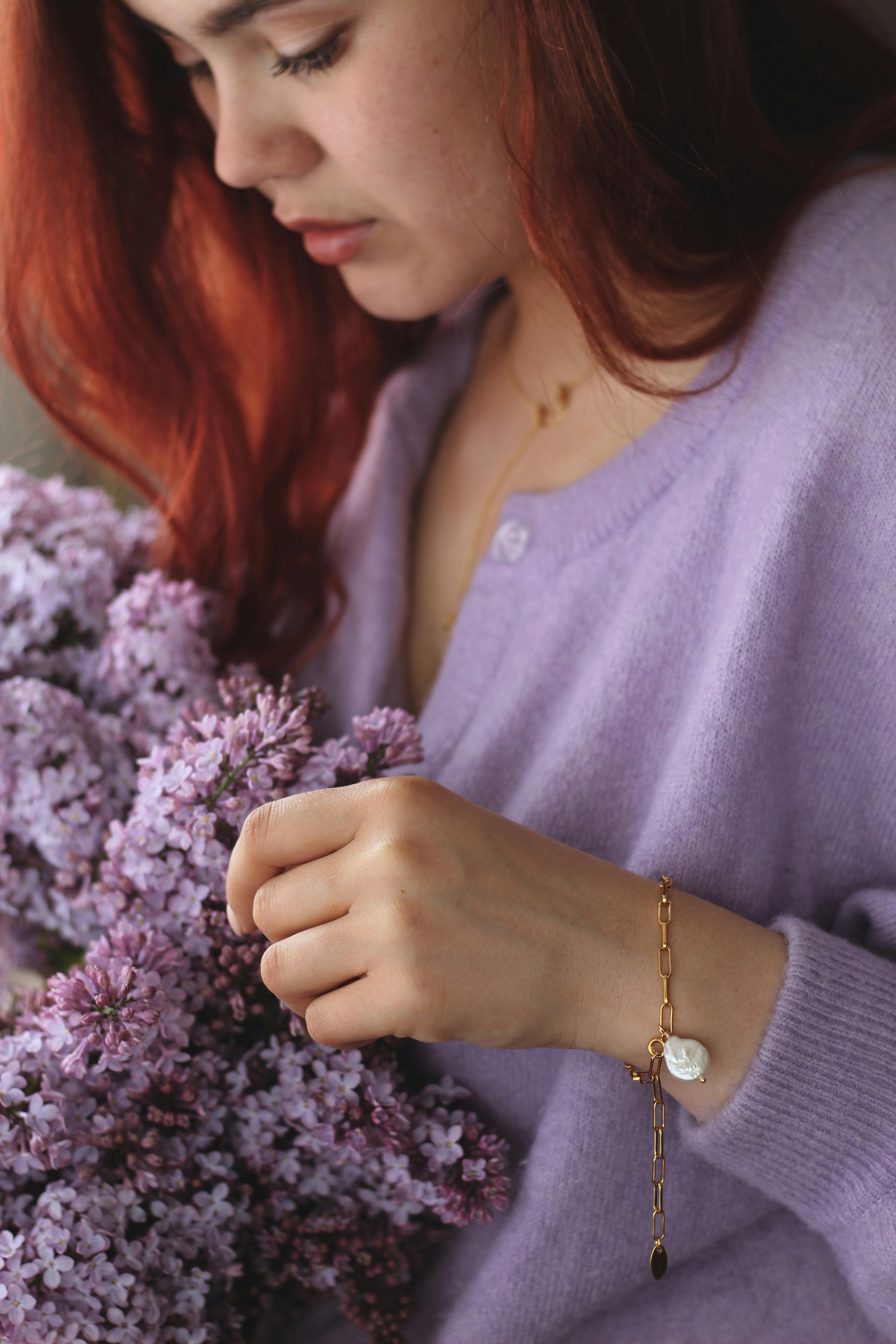 Woman wearing gold bracelet holds lilac flowers while looking down.