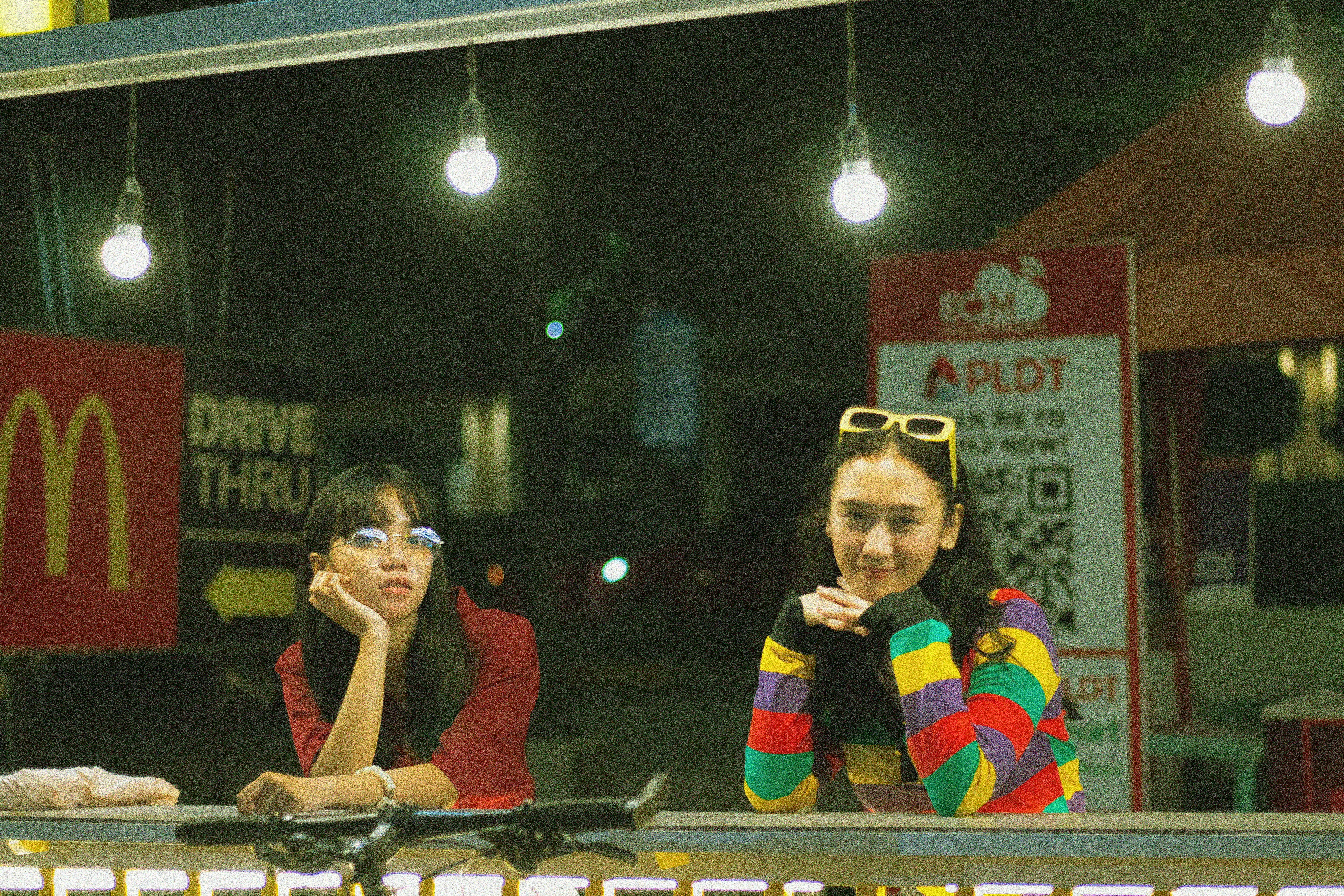 Colorful night scene with two women under lights at a fast-food outlet's drive-thru.