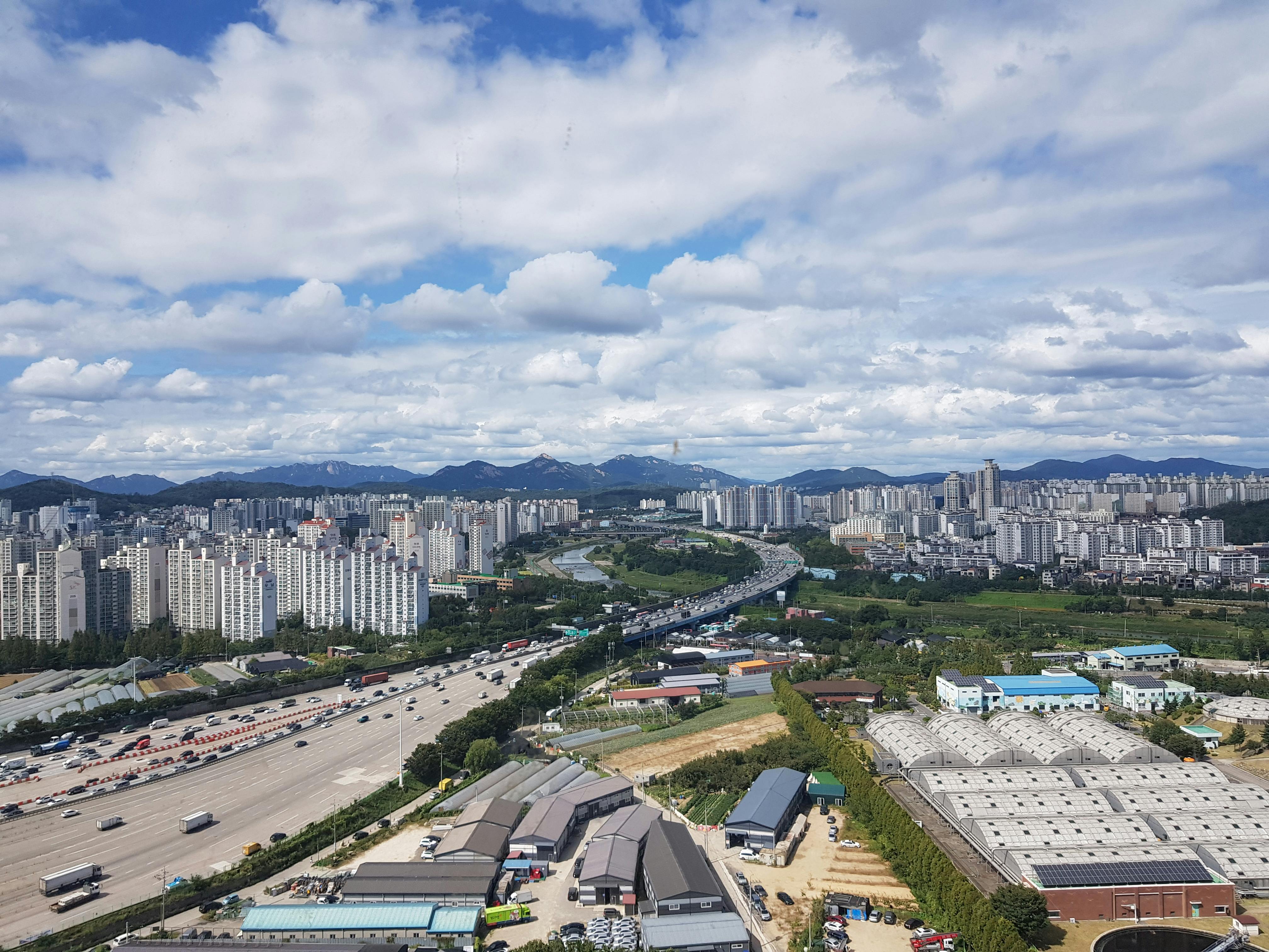 Aerial panorama of Guri City with urban skyline, highways, and mountains under a cloudy sky.