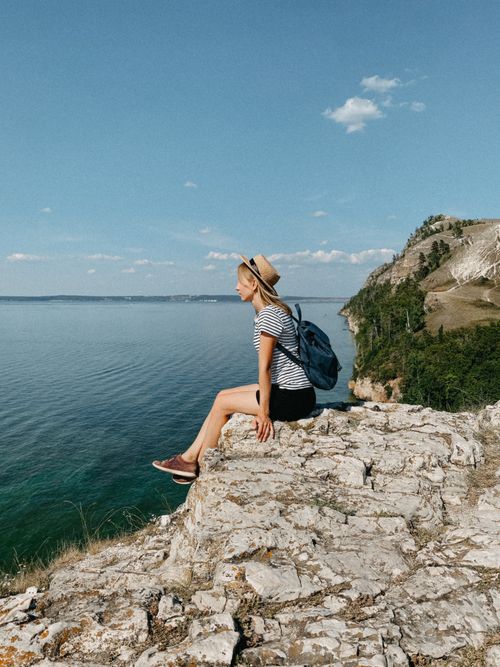 Free A woman wearing a hat sits on a cliff edge, enjoying the ocean view on a sunny day. Stock Photo