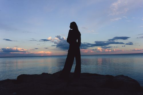 Free A serene silhouette of a woman standing by the ocean during a colorful sunset. Stock Photo