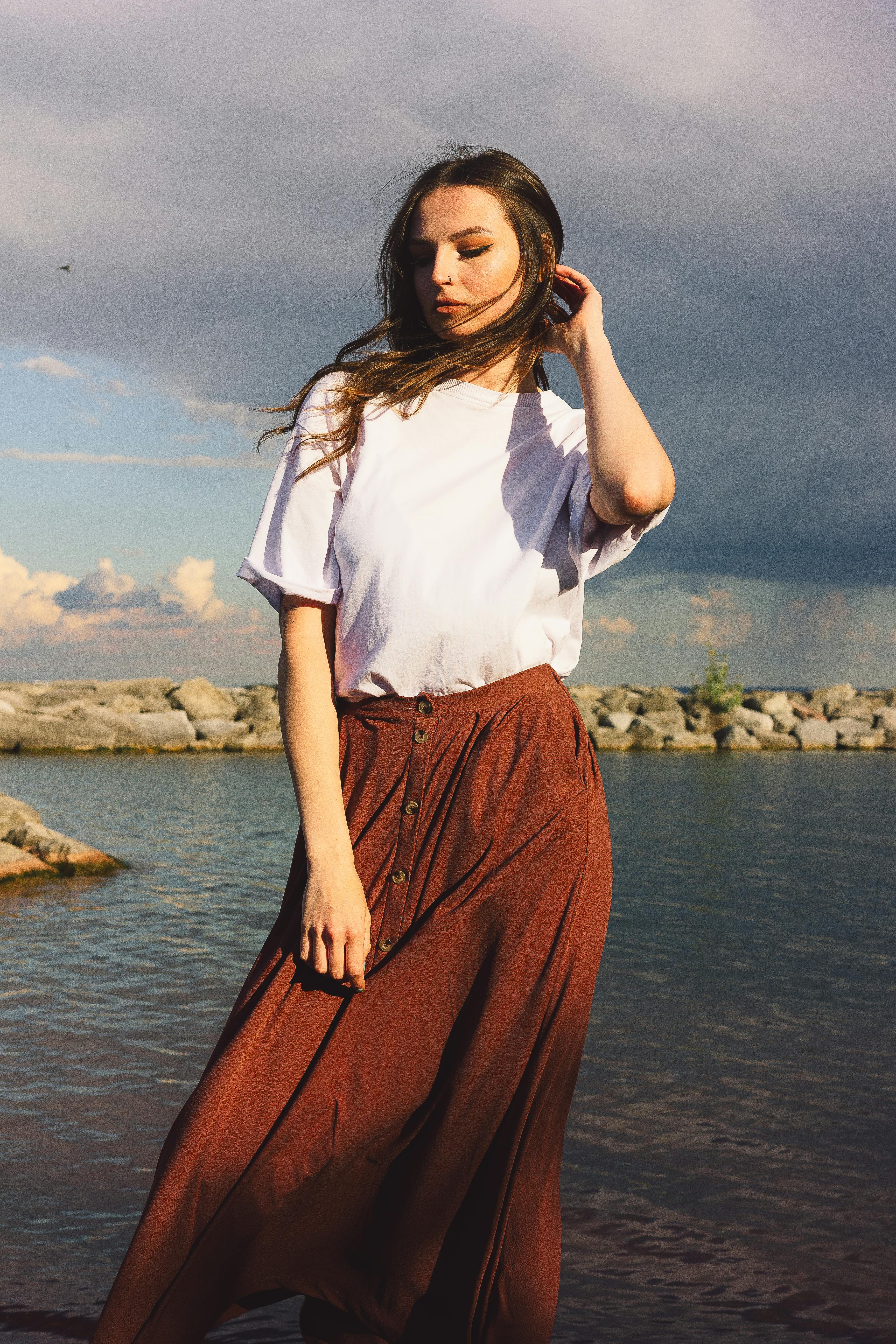 Woman in casual attire standing with serene expression near water's edge.