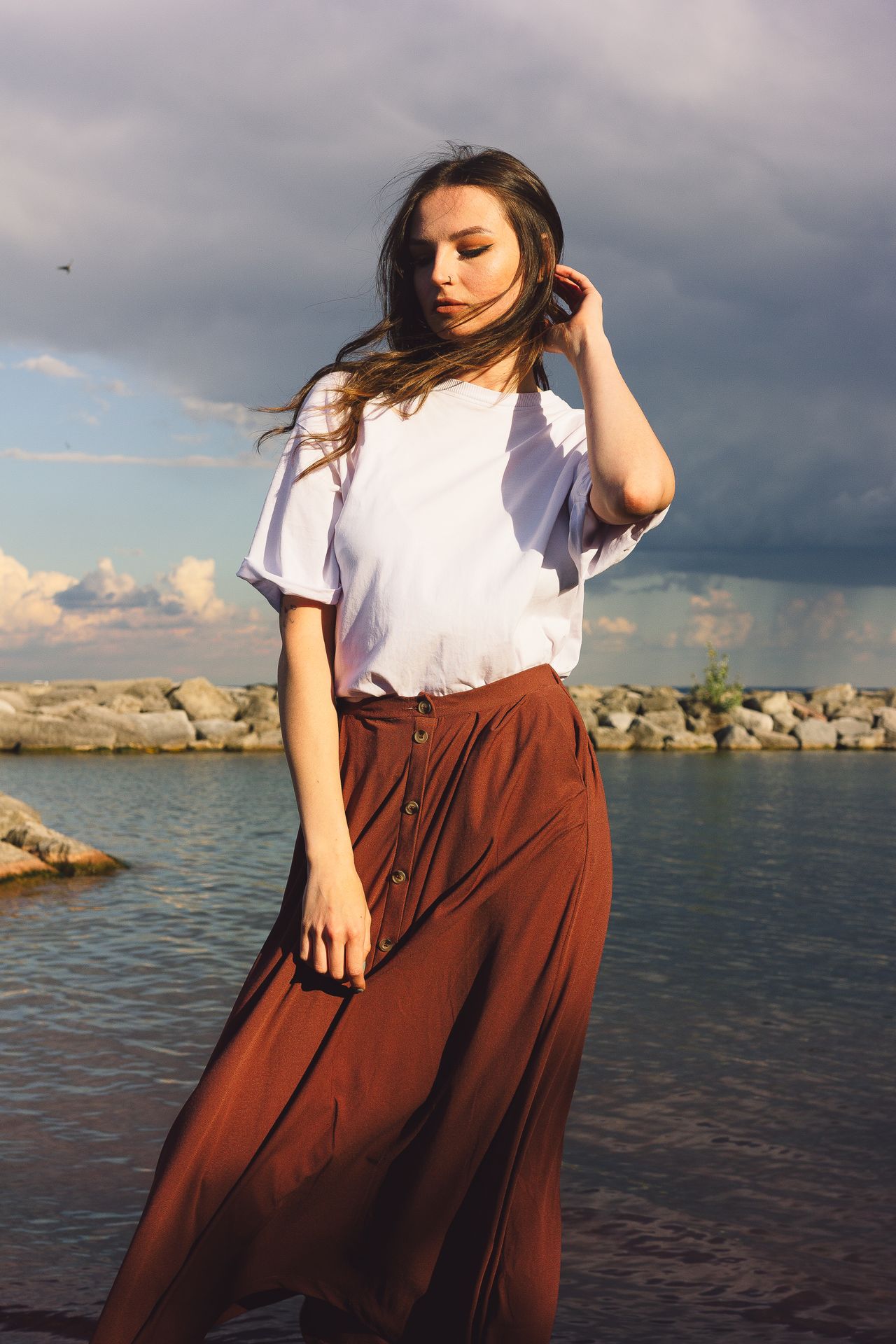 Woman in a white tee and rust button-front midi skirt standing by the water