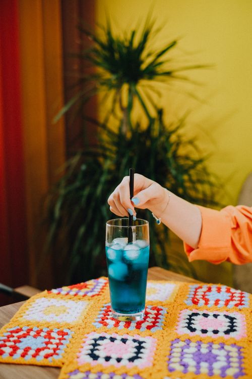 Free A vibrant blue cocktail with ice, stirred by a hand, on a crocheted tablecloth indoors. Stock Photo