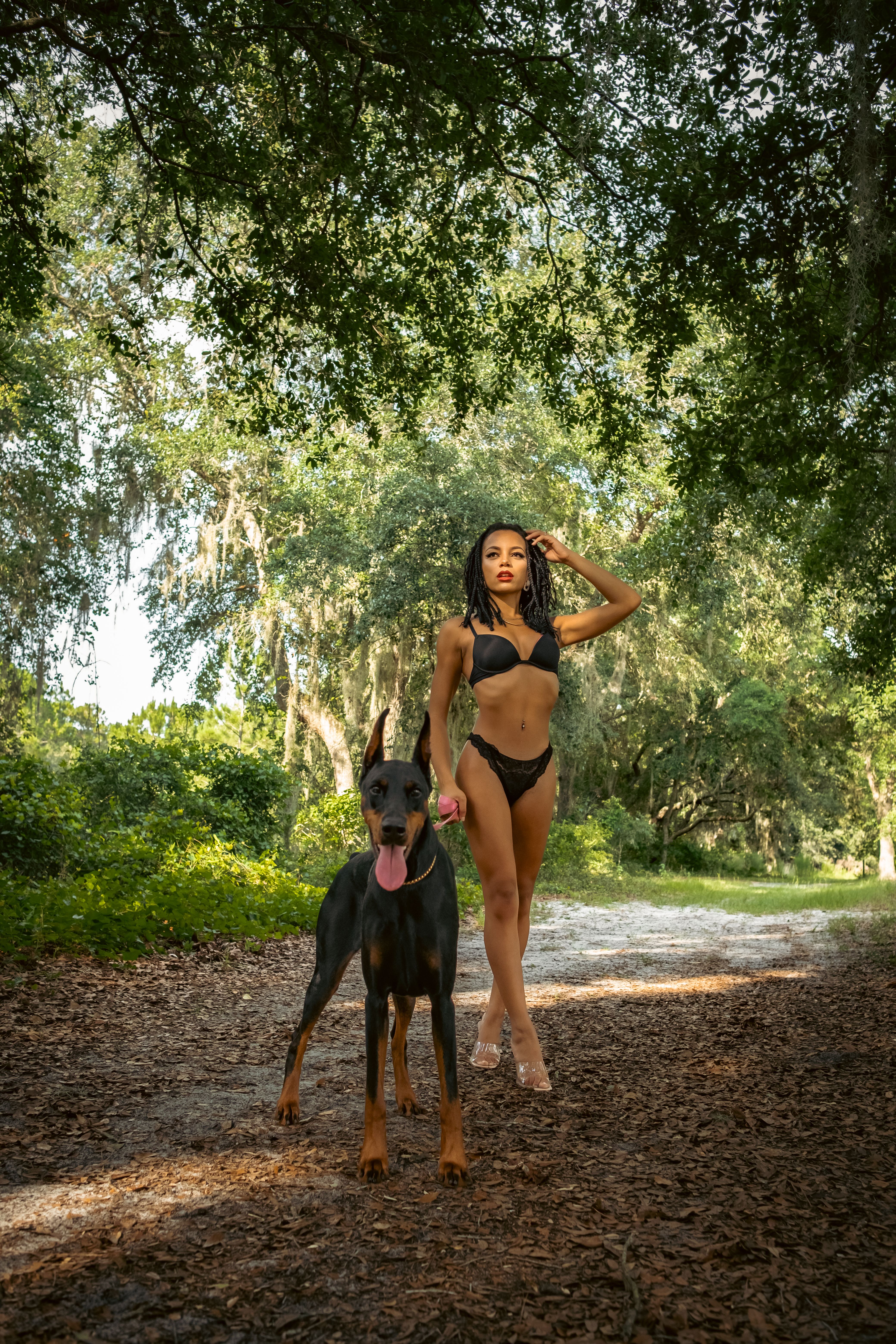 Black woman in lingerie with her Dobermann dog standing on a forest path.