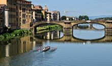 Rowers glide under a historical bridge in Florence, reflected in the river.