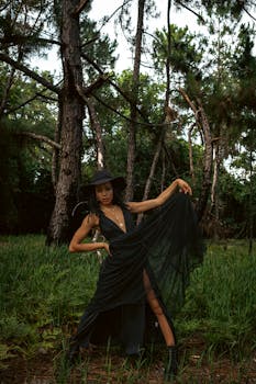 Stylish woman in a black dress and hat poses elegantly in a lush forest setting.