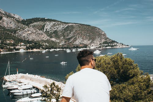 Homem Vestindo Camisa Branca E óculos De Sol Na Frente De Barcos E Montanha Enquanto Olha Para O Lado Direito Sob O Céu Azul