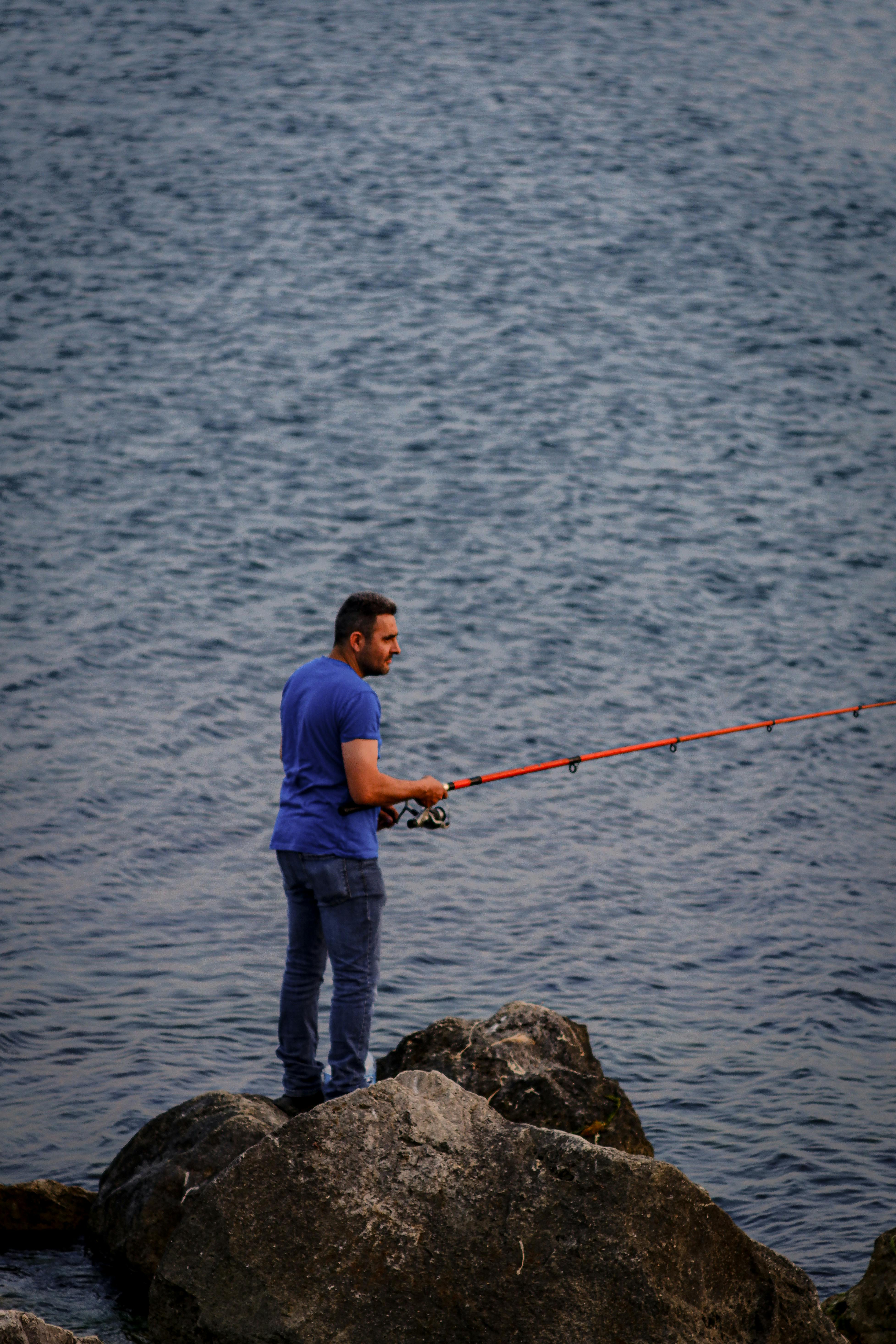 a man in blue shirt standing on the rock while holding a fishing rod