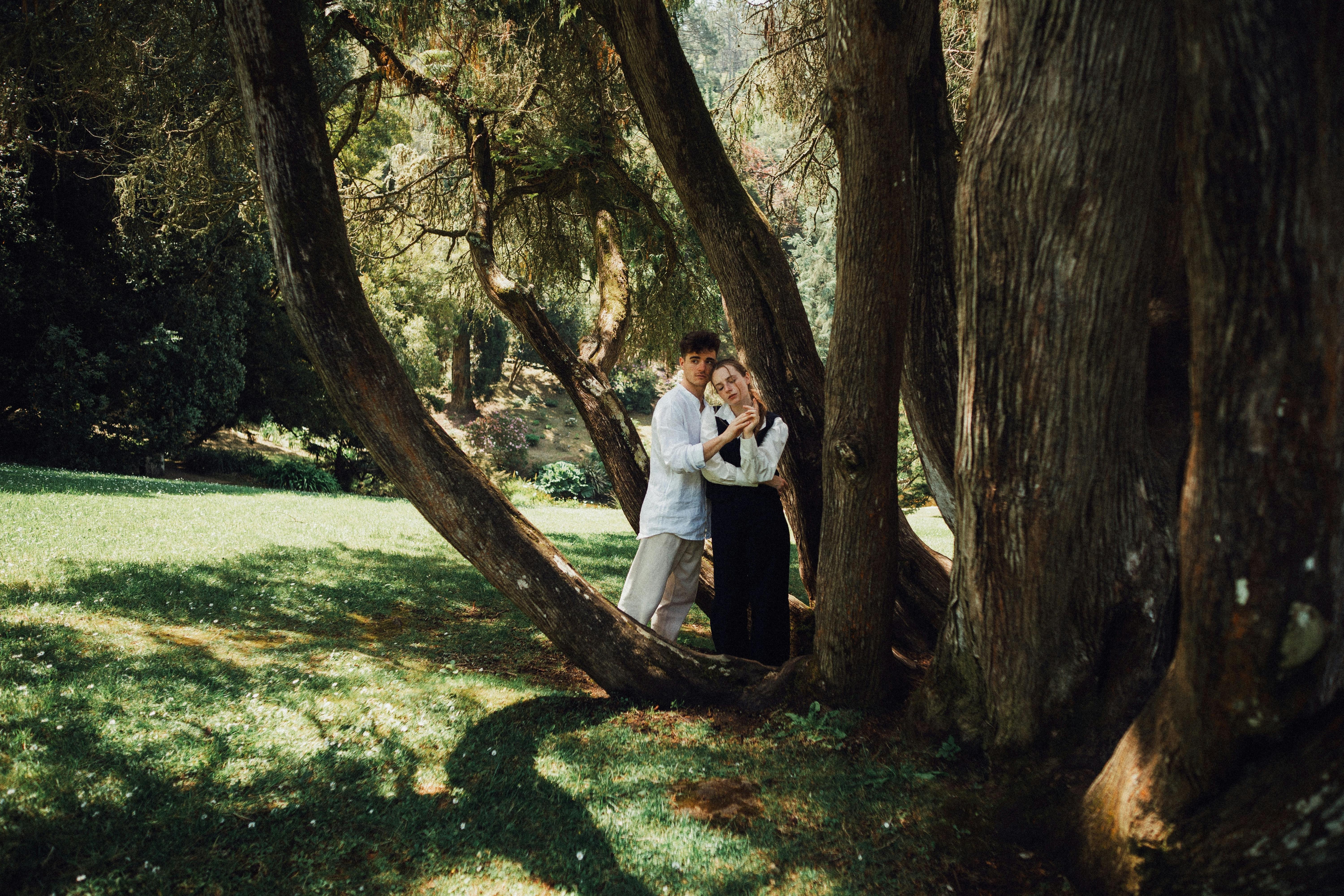 A couple embracing under large trees in a lush forest park setting, capturing serenity and nature.