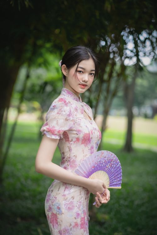 Free Beautiful Asian woman in floral traditional dress holding a fan in a serene outdoor setting. Stock Photo