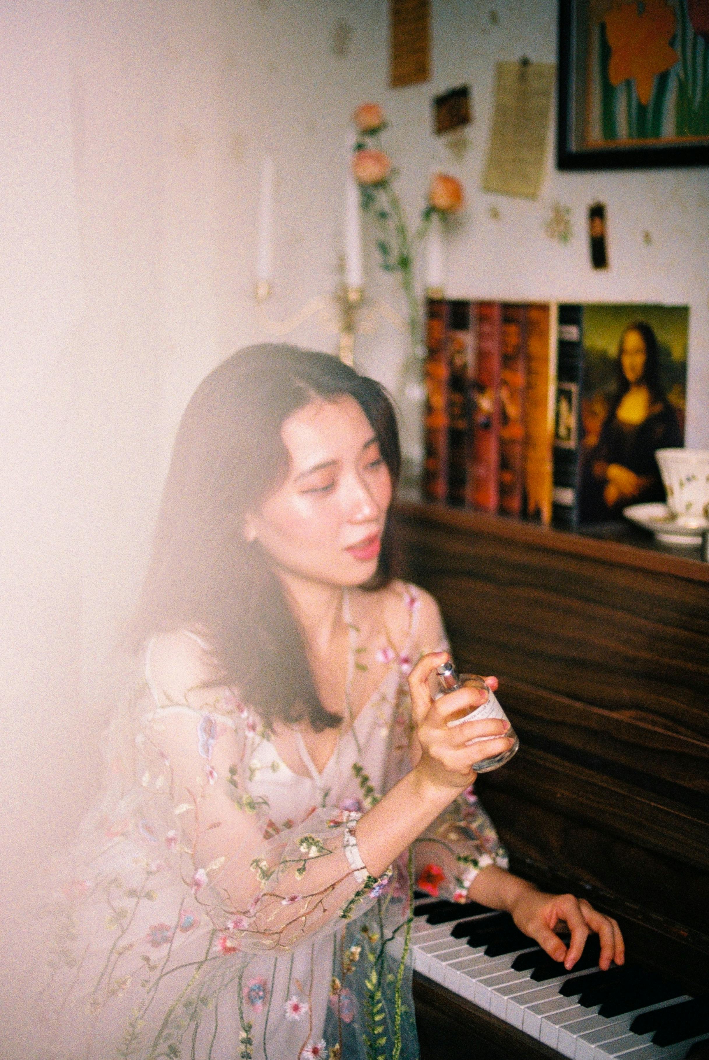 Asian woman in a floral dress spraying perfume while sitting at a piano indoors with a soft artistic flare.
