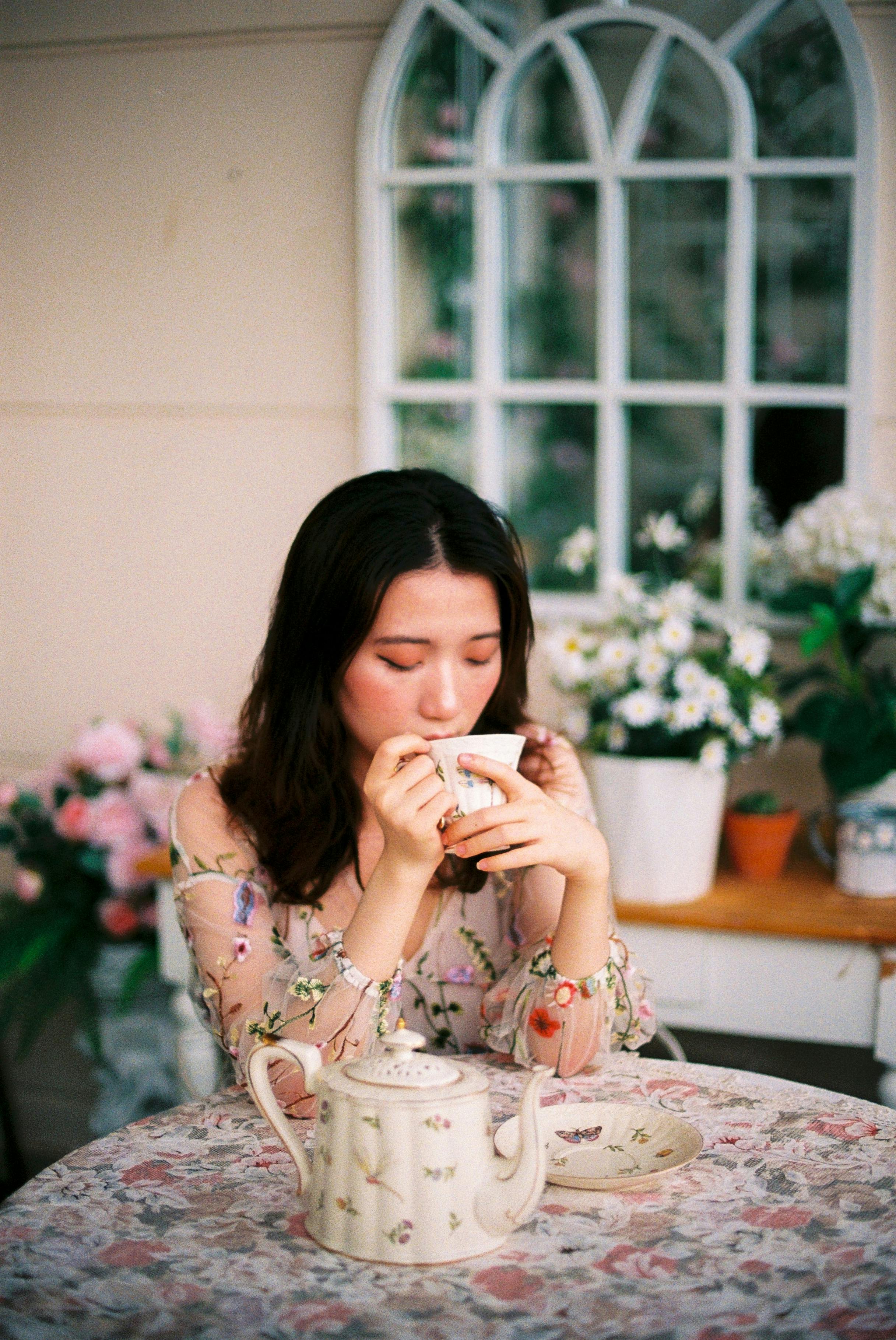 Woman savoring tea at a floral table with a serene expression, surrounded by flowers.