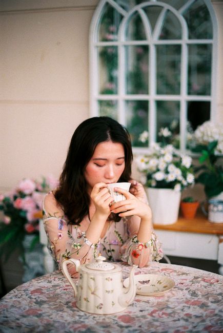 Woman savoring tea at a floral table with a serene expression, surrounded by flowers.