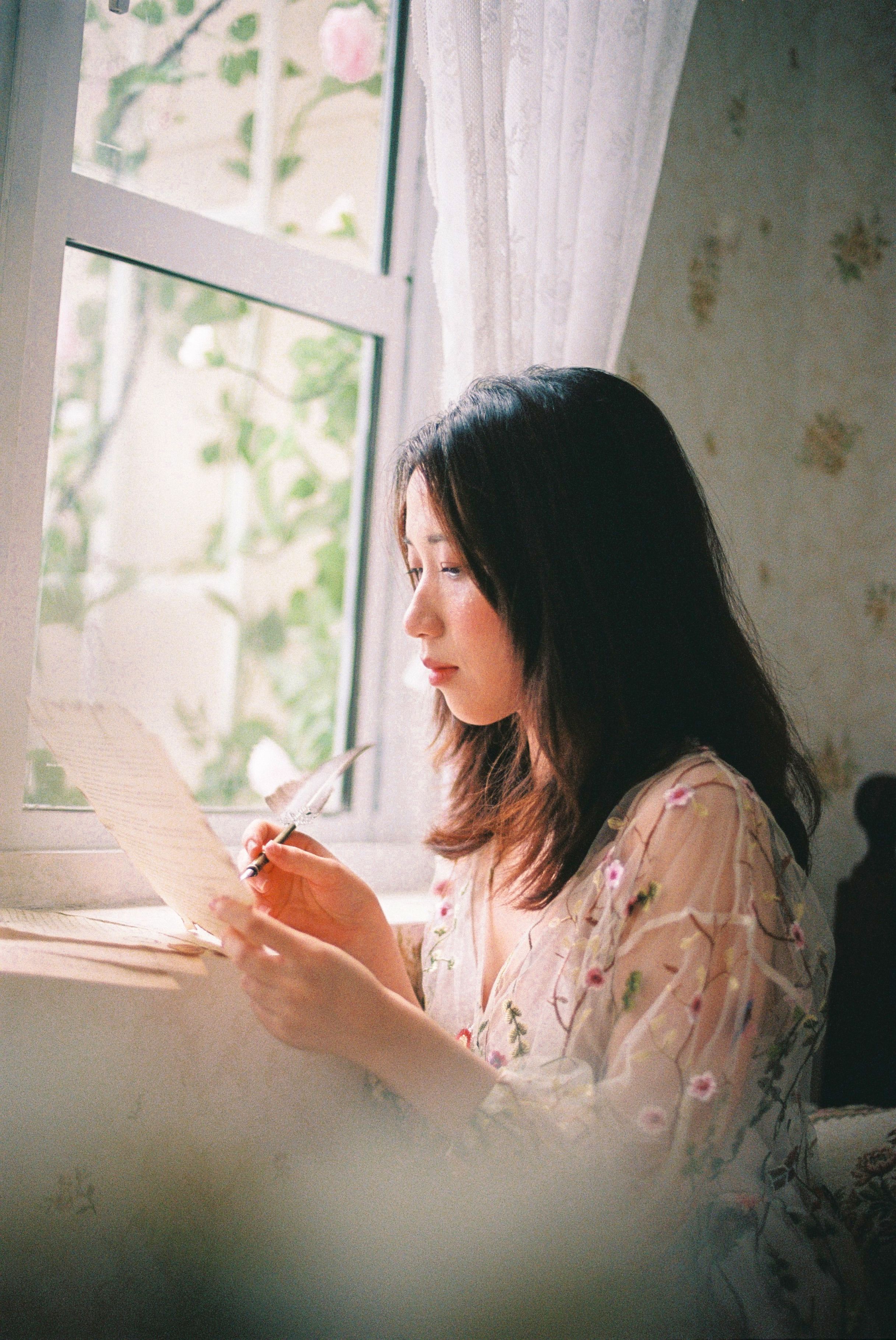 A woman in a floral lace top writes by a sunlit window, using a quill pen.