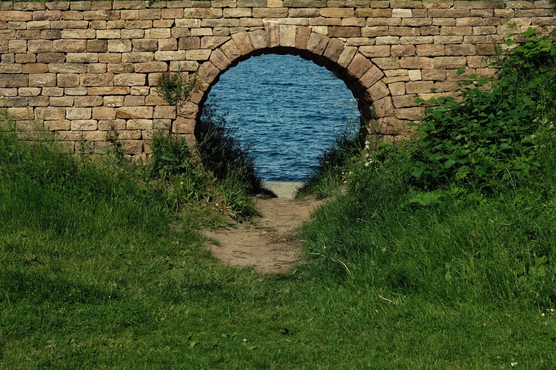 Free A serene view through a stone archway revealing a tranquil body of water surrounded by lush greenery. Stock Photo