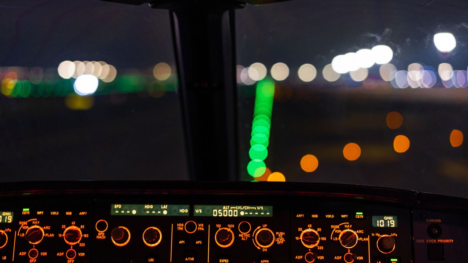Close-up of an aircraft cockpit dashboard illuminated at night with bokeh runway lights.