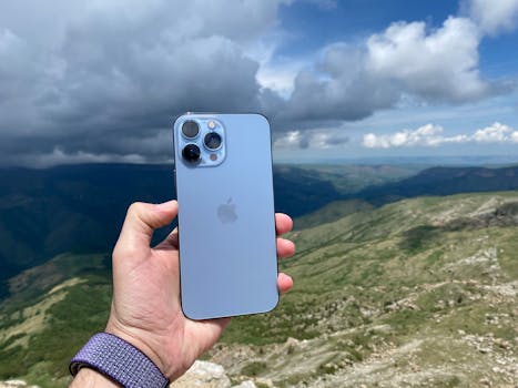 A hand holding a smartphone against a scenic mountain landscape with a cloudy sky.