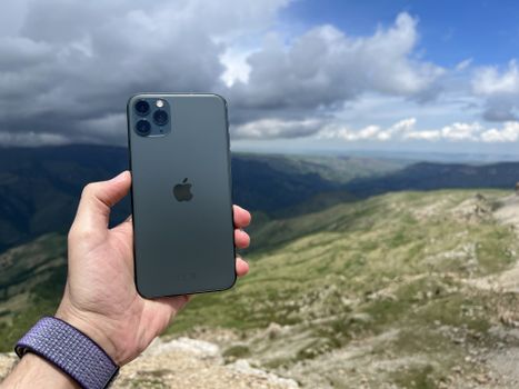 A hand holding a smartphone with three cameras, overlooking a scenic mountainous landscape.