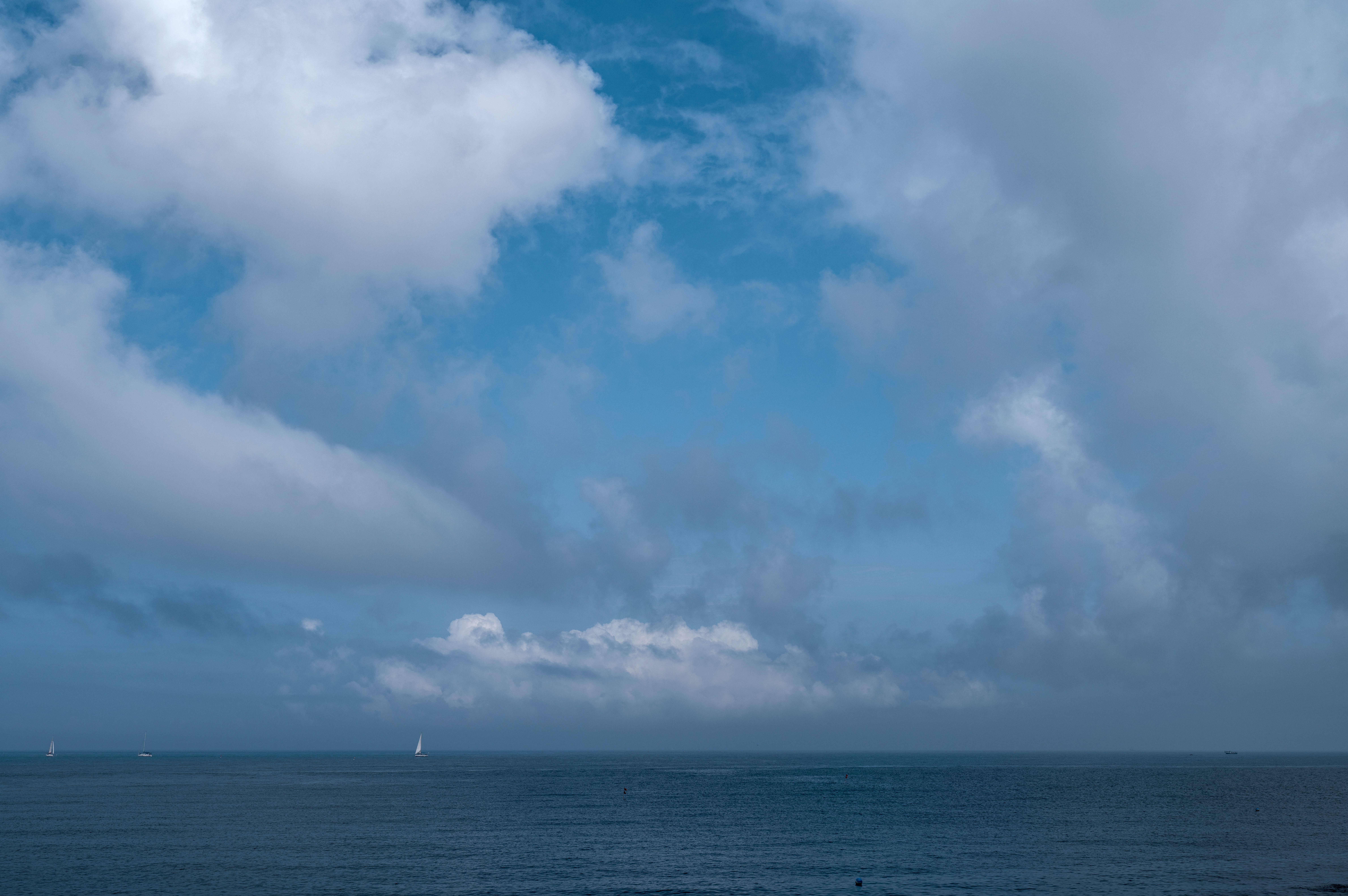 Calm seascape with sailboats and dramatic clouds under a bright blue sky.