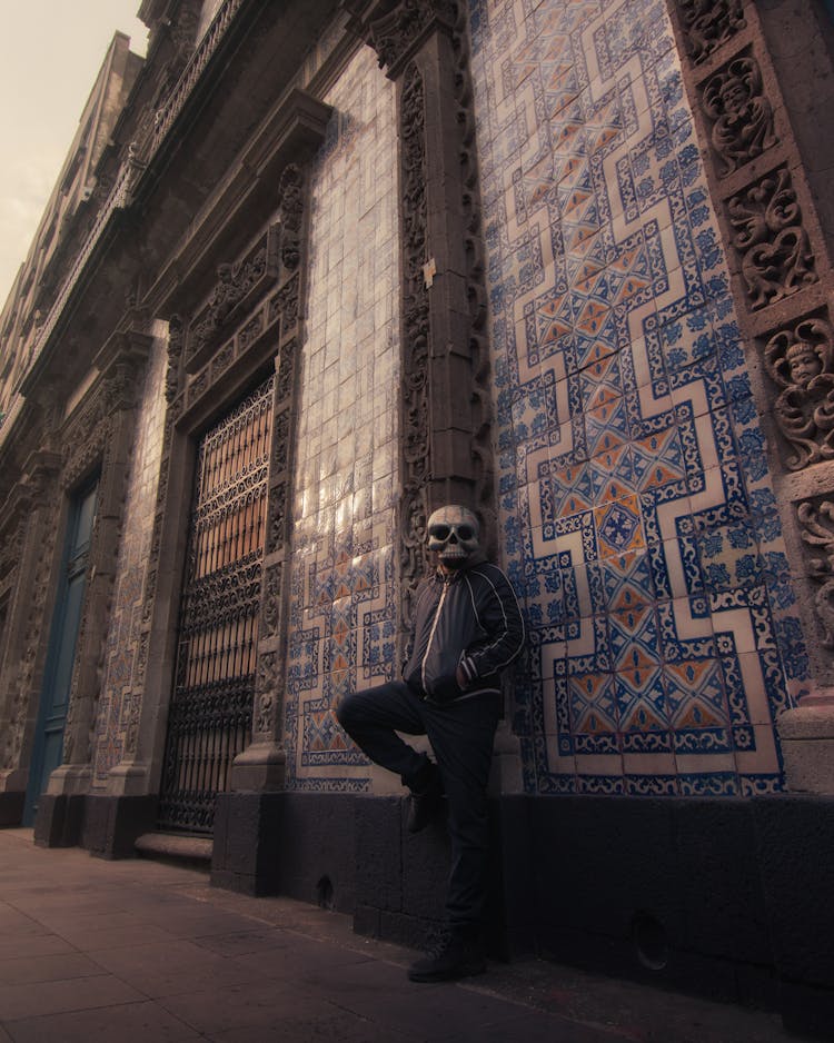 Man In A Skull Mask Standing Against A Wall Of A Building With An Artistic Pattern 