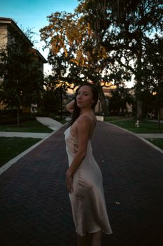 Elegant woman in satin white dress standing on a brick pathway at dusk. Soft lighting creates a serene atmosphere.