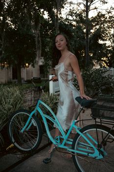 A woman in a satin dress stands gracefully next to a turquoise bicycle at sunset.