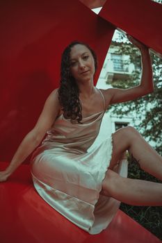 A woman in a satin dress elegantly poses on a large red structure outdoors.