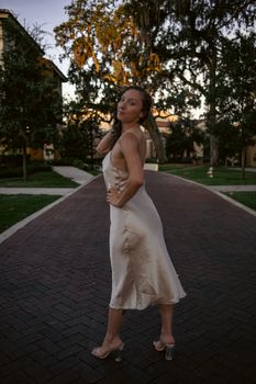 Woman posing in a satin dress on a dimly lit stone pathway lined with trees.