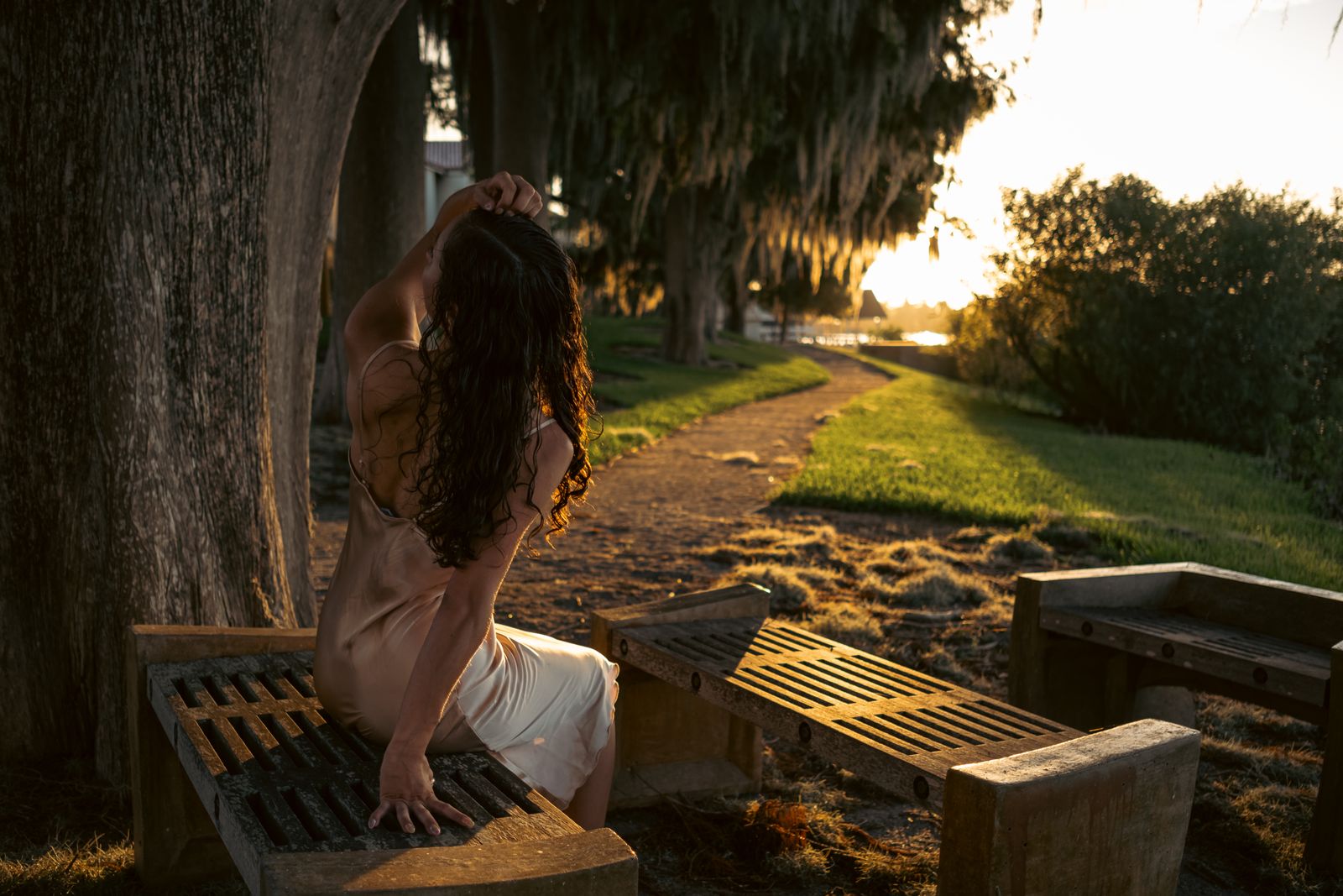 Woman in a satin slip dress sits on a wooden bench, touching her long dark hair at sunset