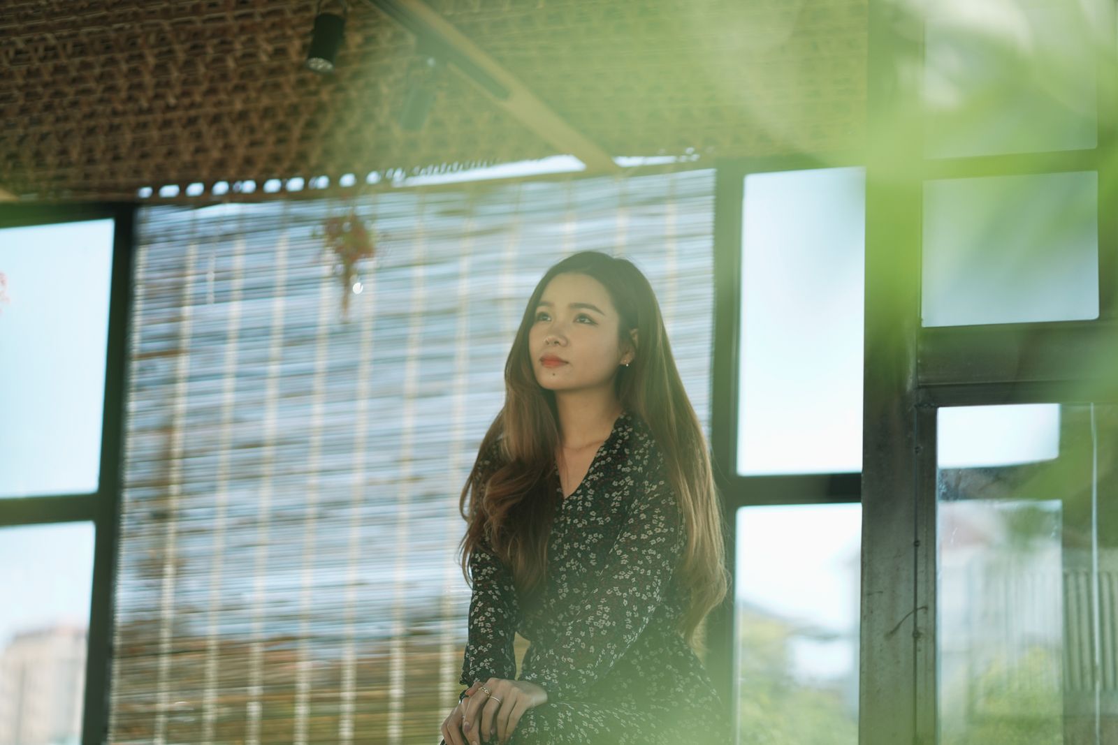 Woman with long brown hair sitting by sunlit windows, blurred greenery in the foreground