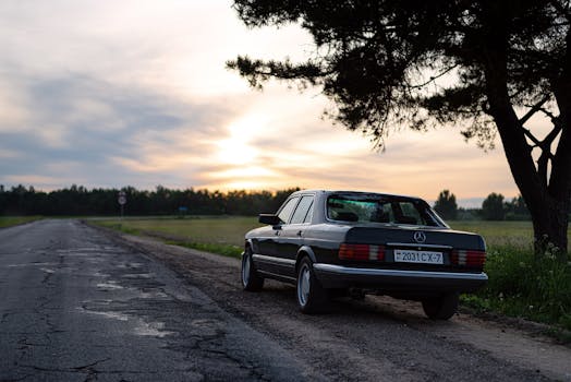 A classic Mercedes Benz parked by a rural road during a scenic sunset in a serene landscape.