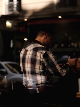 Barber wearing plaid shirt styling a customer's hair in an urban setting.