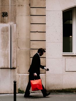 Elegant businessman walking on sidewalk with striking red bag against urban building exterior.