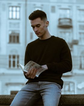 A young man sits on a ledge reading a book against an urban backdrop, offering a moment of quiet amidst city life.