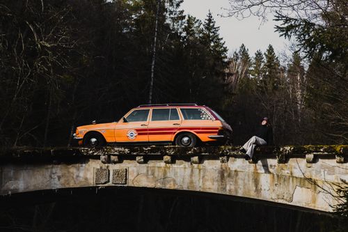 Free A classic orange car parked on a rustic forest bridge with a man relaxing beside it. Stock Photo
