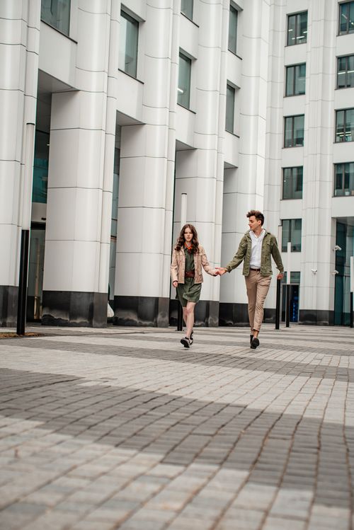 Free A young couple holding hands while walking past modern buildings in a city setting. Stock Photo