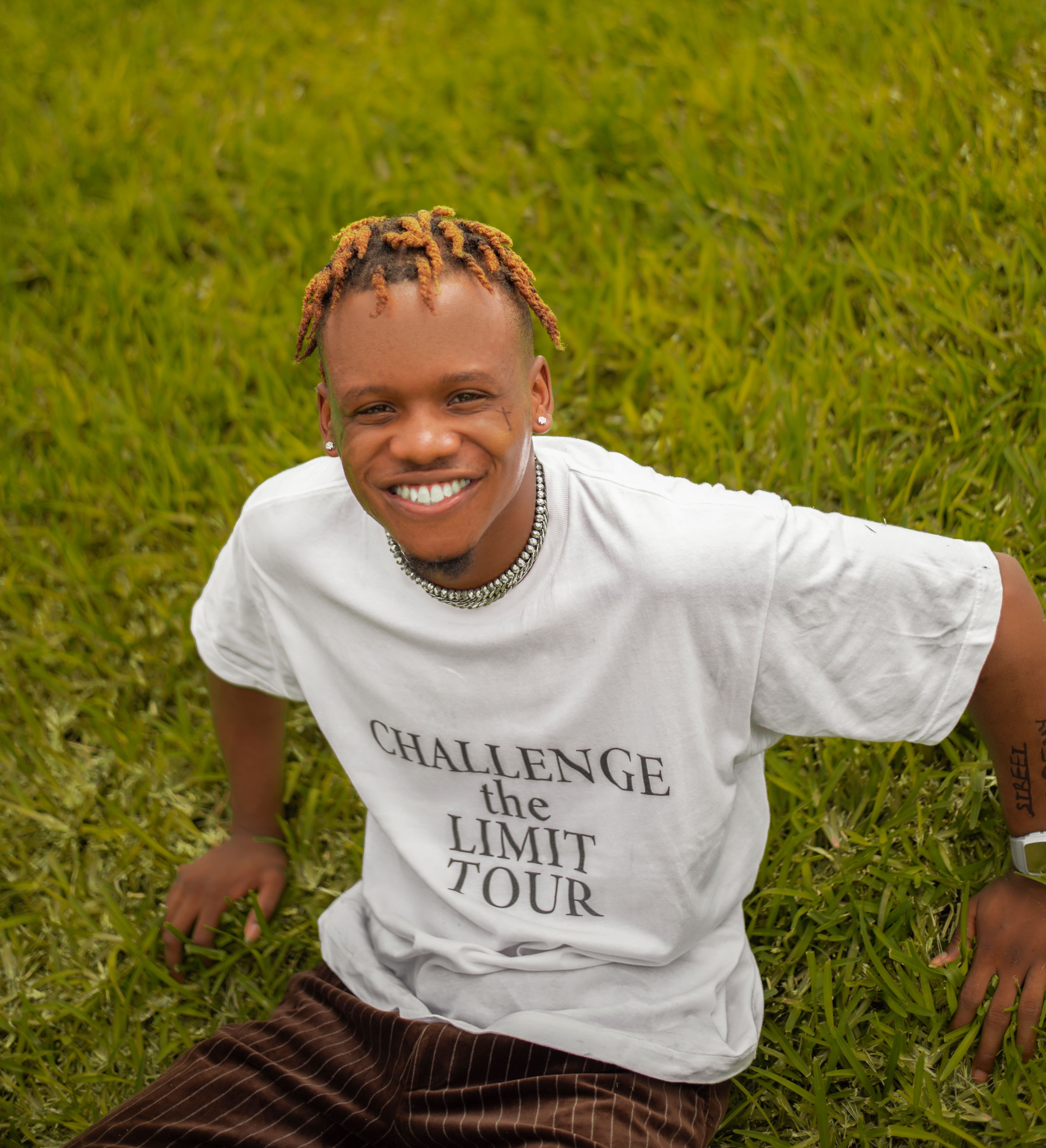 Cheerful man sitting on grass, wearing 'Challenge the Limit Tour' shirt.