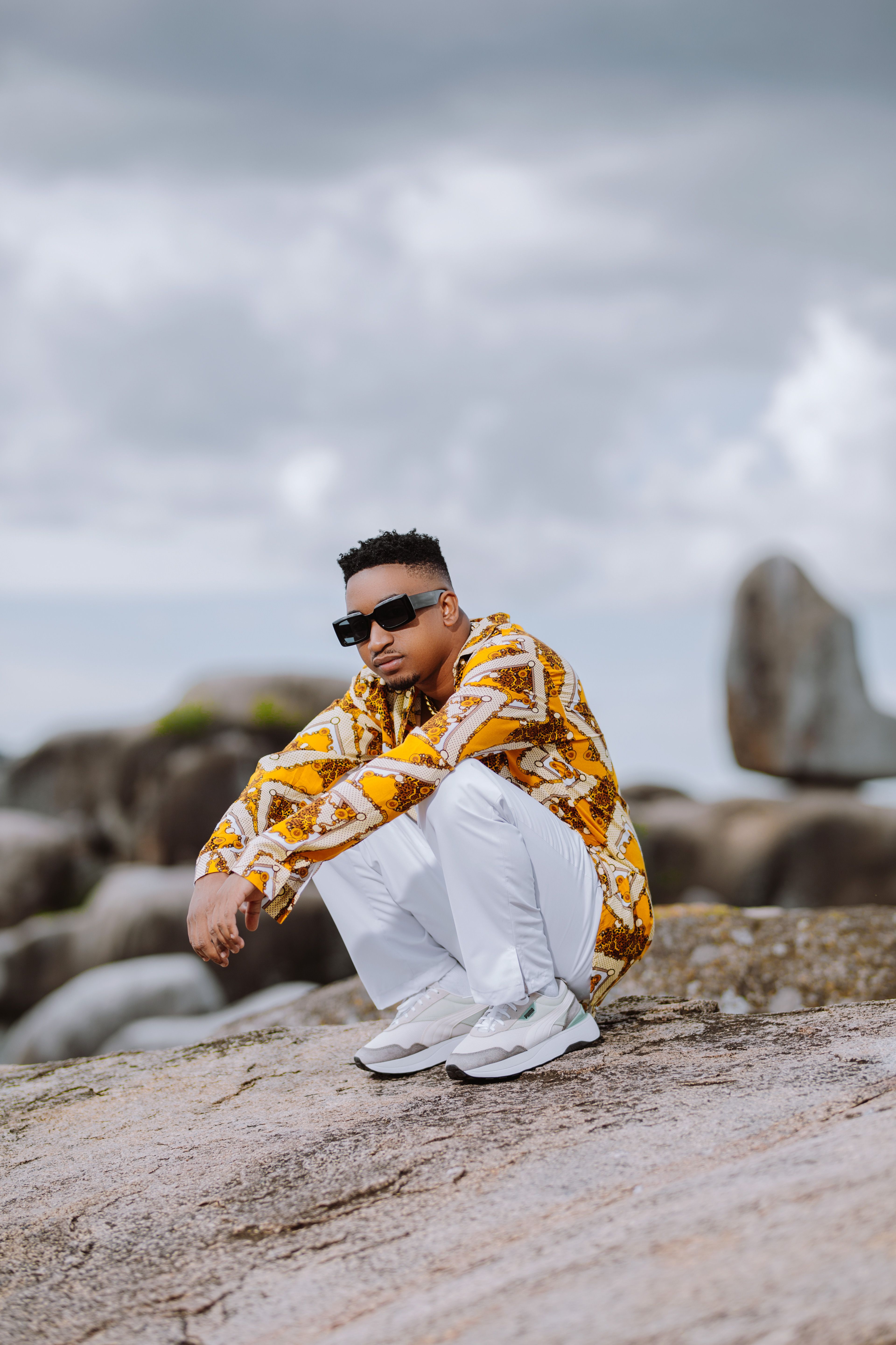 Fashionable man in sunglasses crouching on rocks, wearing a vibrant shirt.