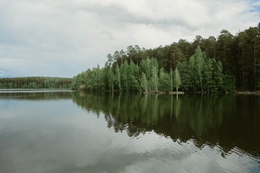 Calm lake reflecting lush green trees under cloudy sky, showcasing natural beauty.