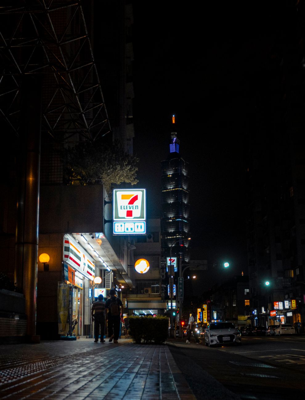 Vibrant night scene of a downtown street featuring a well-lit skyscraper and bustling urban life.