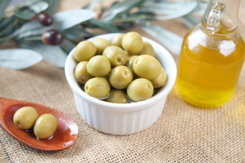 White ramekin of green olives beside a glass bottle of olive oil on burlap